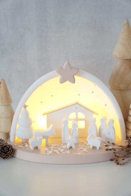 Lichtdurchflutete Weihnachtskrippe mit weißen Figuren und Tannenbäumen im Hintergrund.